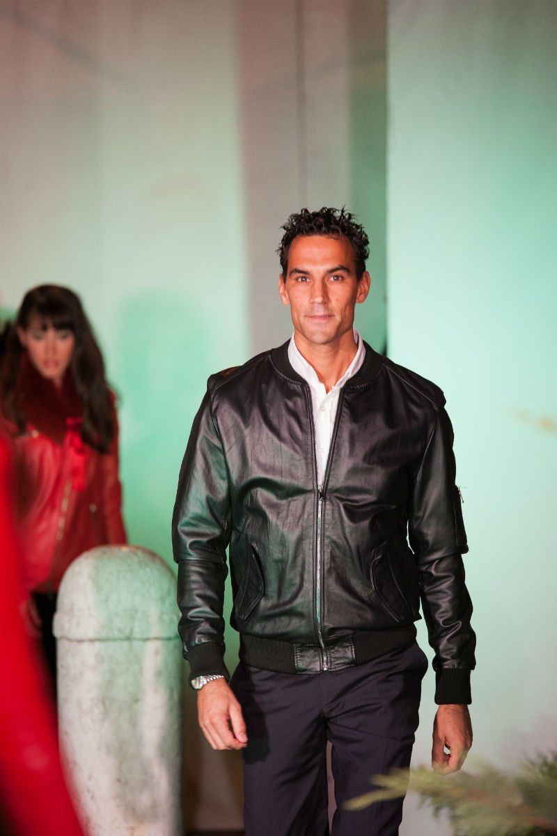 Uomo con giacca di pelle nera e camicia bianca, che cammina; donna con giacca rossa sullo sfondo.