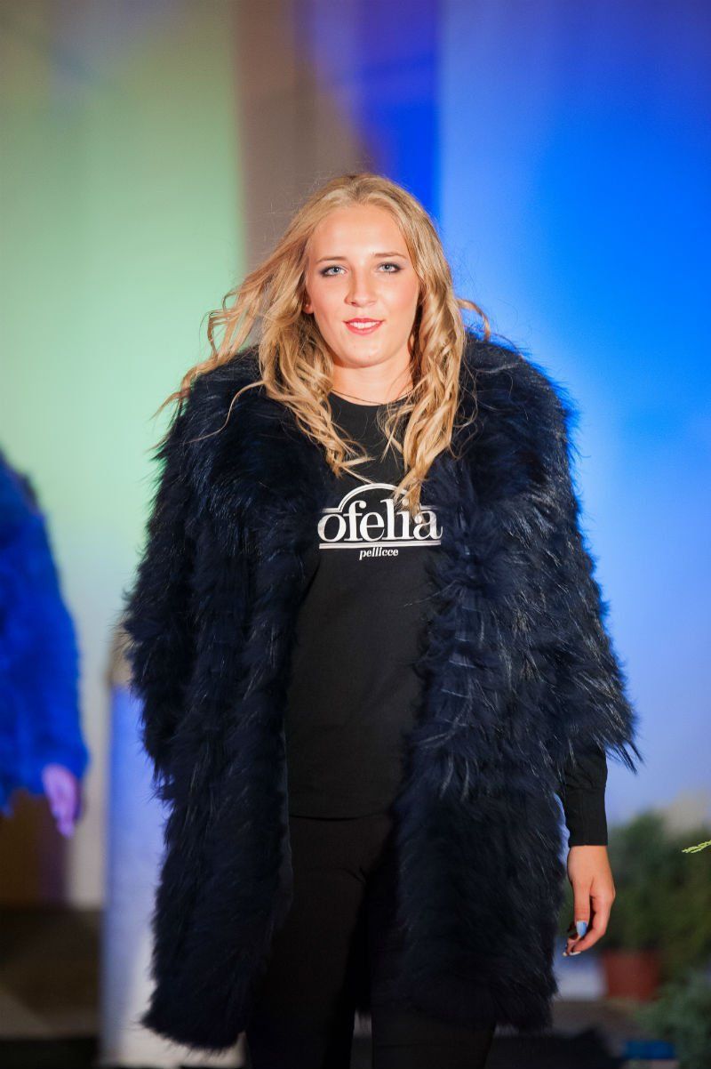 Modella bionda con pelliccia blu navy e abito nero su una passerella, davanti a uno sfondo blu.