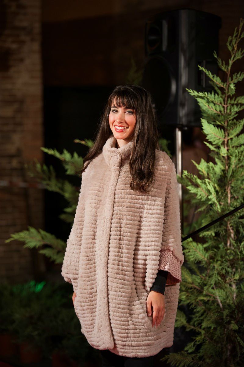 Donna sorridente con cappotto marrone chiaro in piedi in mezzo al verde, probabilmente in occasione di un evento o di una mostra.