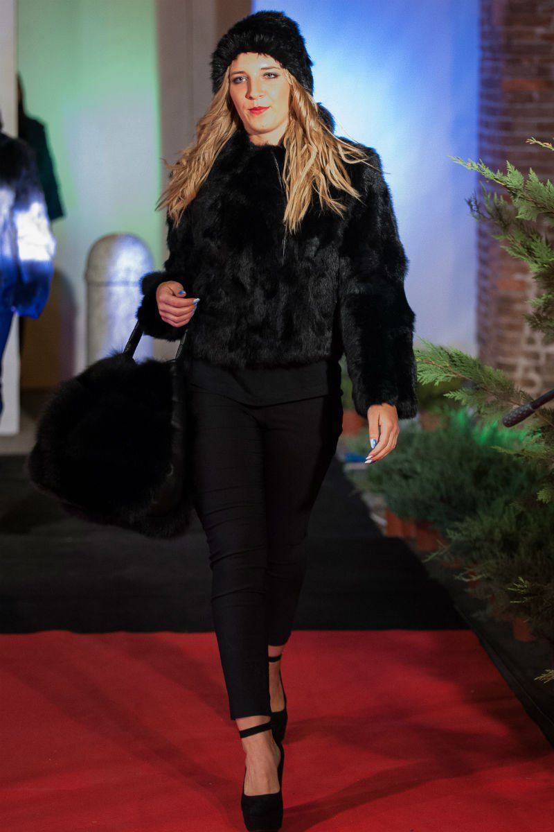 Una donna con un cappotto di pelliccia nero e un cappello cammina sul red carpet, tenendo in mano una borsa, durante una sfilata di moda.