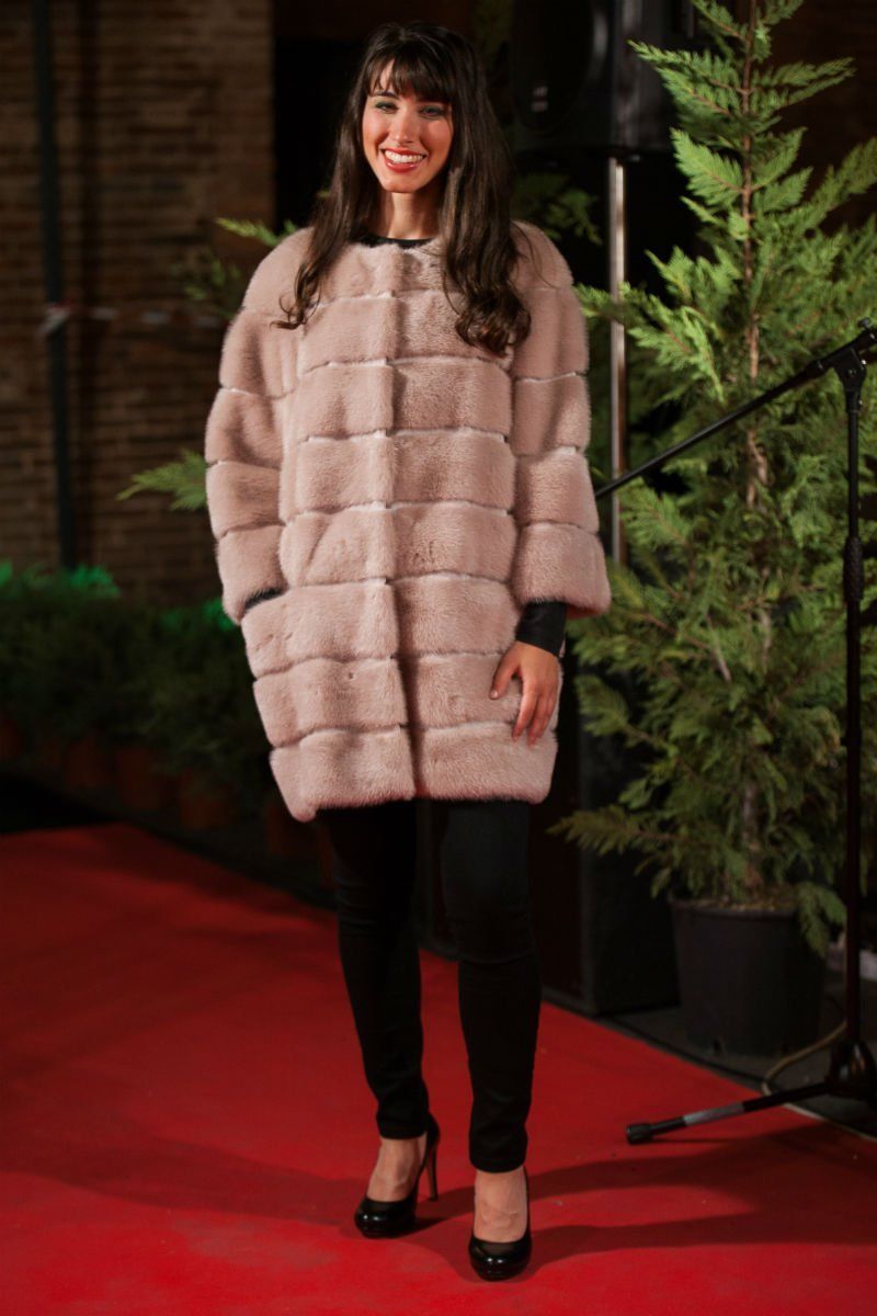 Una donna che indossa un cappotto di pelliccia marrone chiaro, pantaloni neri e tacchi è in piedi su un tappeto rosso vicino al verde.