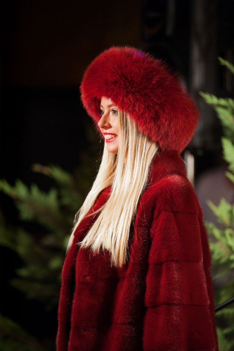 Donna sorridente che indossa un cappello di pelliccia rossa e un cappotto, su uno sfondo scuro.