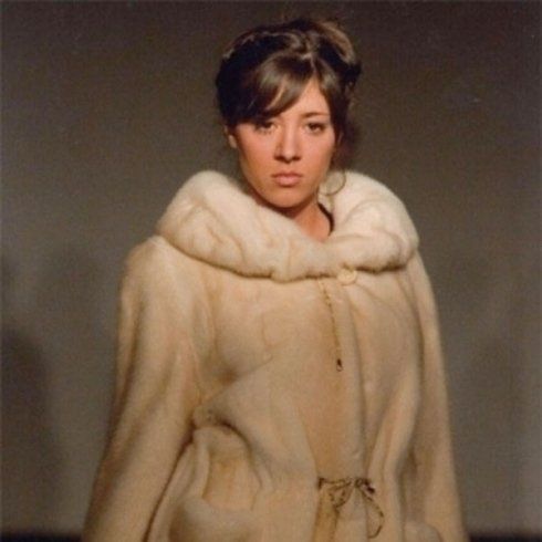 Donna con pelliccia bianca; capelli castani, indossa un top marrone; sfondo neutro.