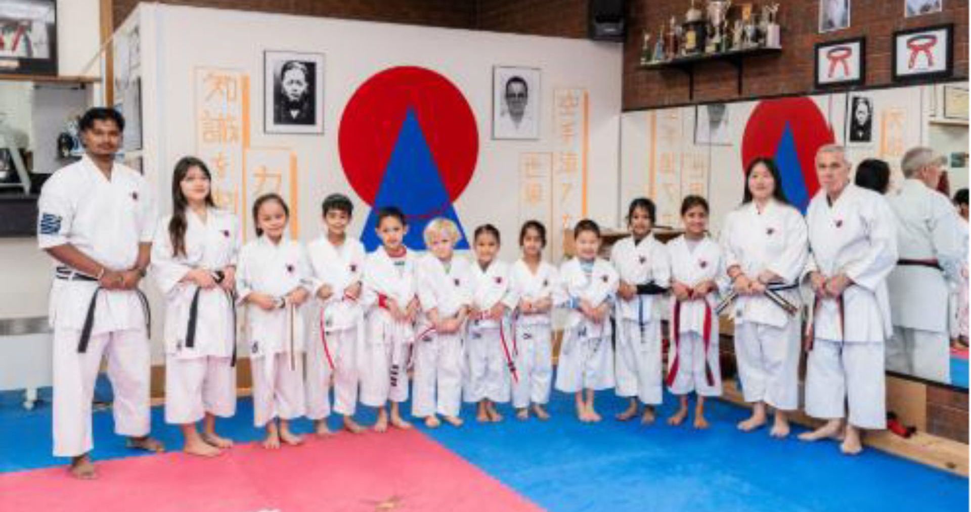 Group of karate students and instructors posing together in a dojo wearing white uniforms and belts.
