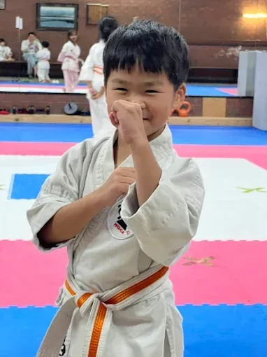 Young boy in a karate uniform, orange belt, smiling and making a fist. Karate class setting.
