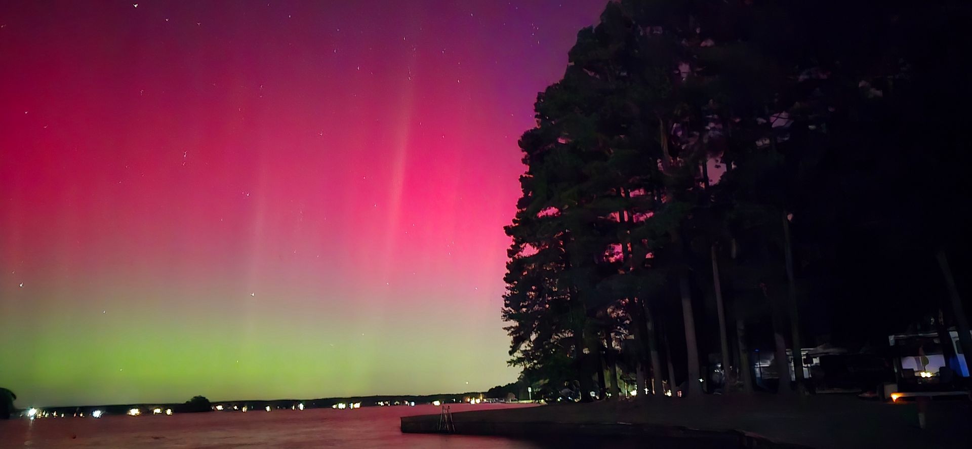 Aurora borealis in a vibrant display over a lake, with trees in silhouette along the shore.