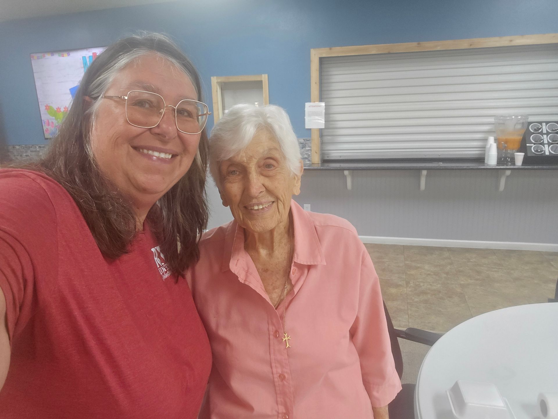 Two smiling people stand together indoors, one in a red shirt and one in a pink shirt.