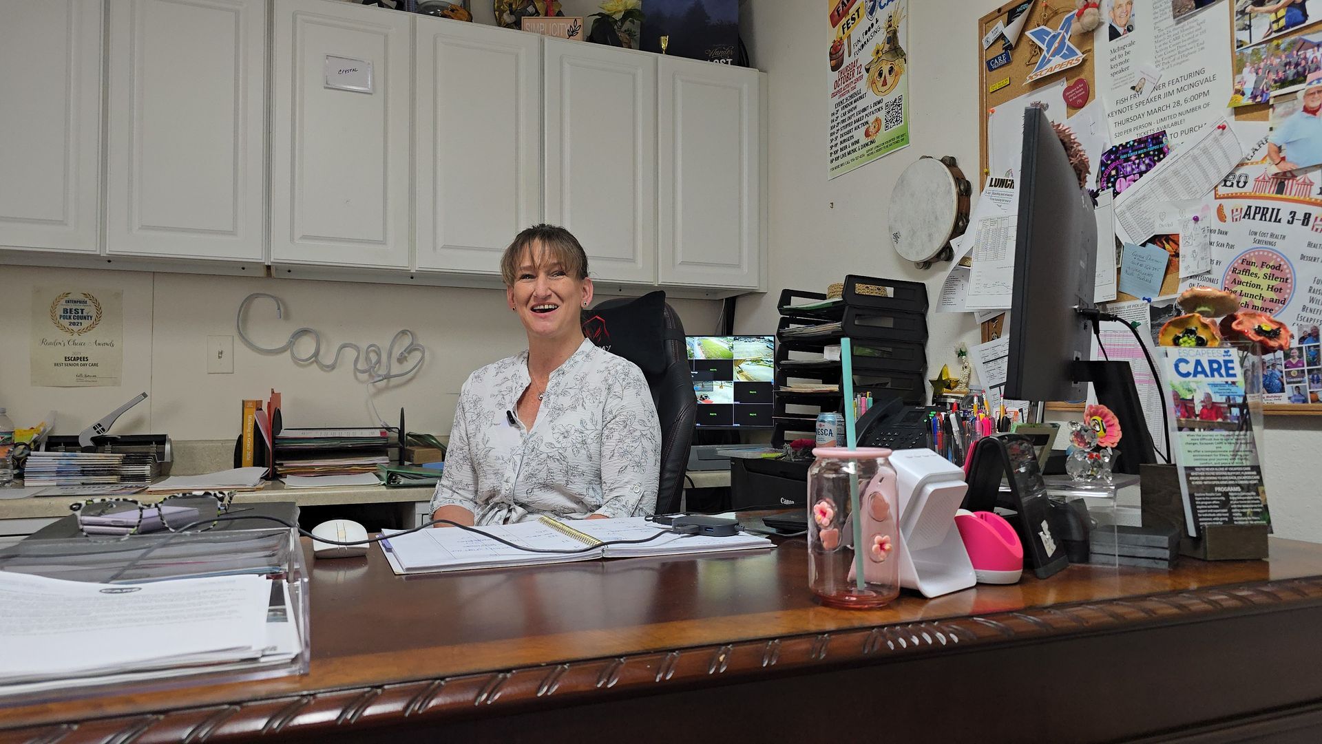 Woman sitting at a cluttered office desk, smiling in a bright room with cabinets and papers.