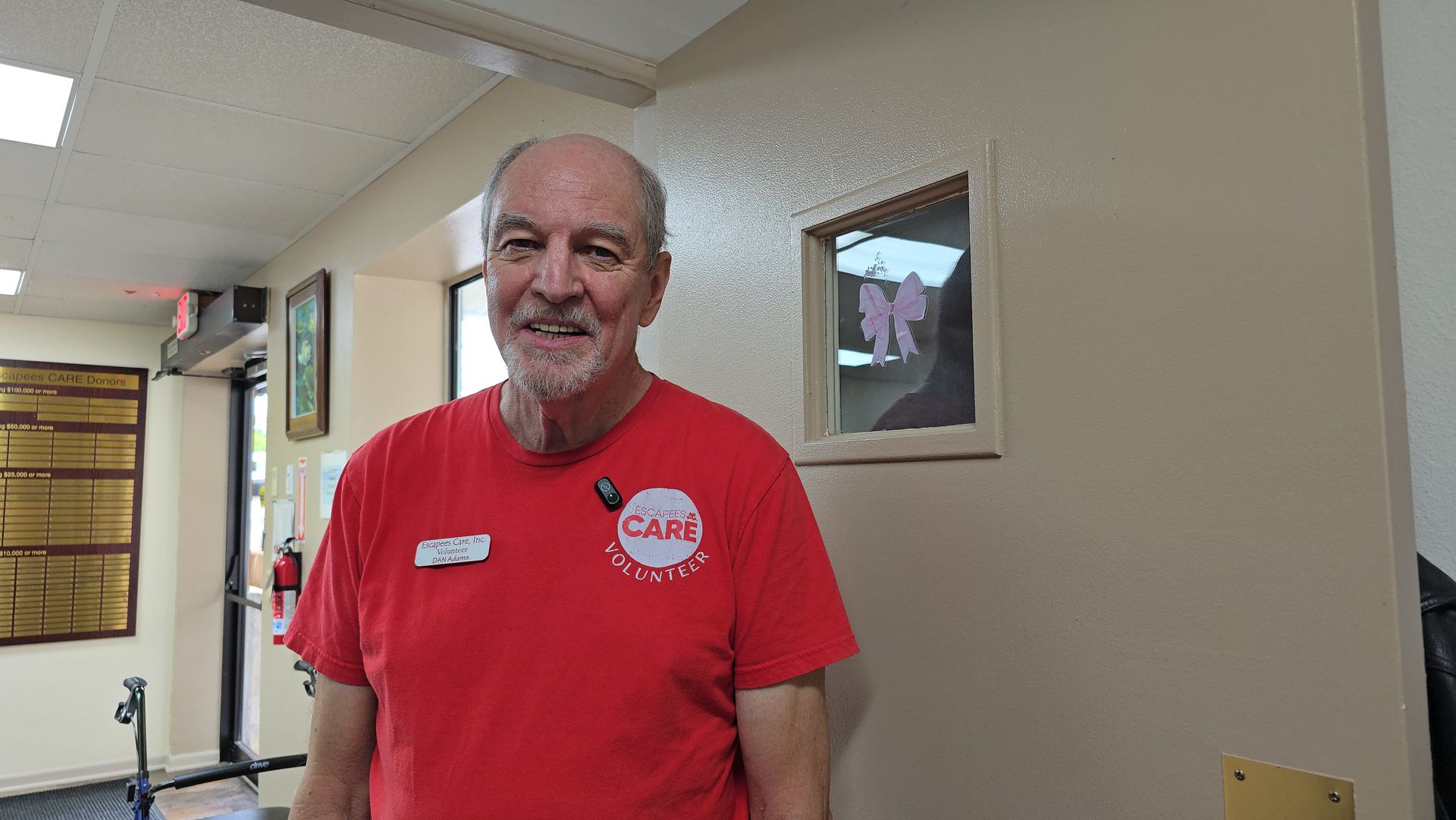 Smiling man in a red shirt standing in a hallway beside framed wall art.