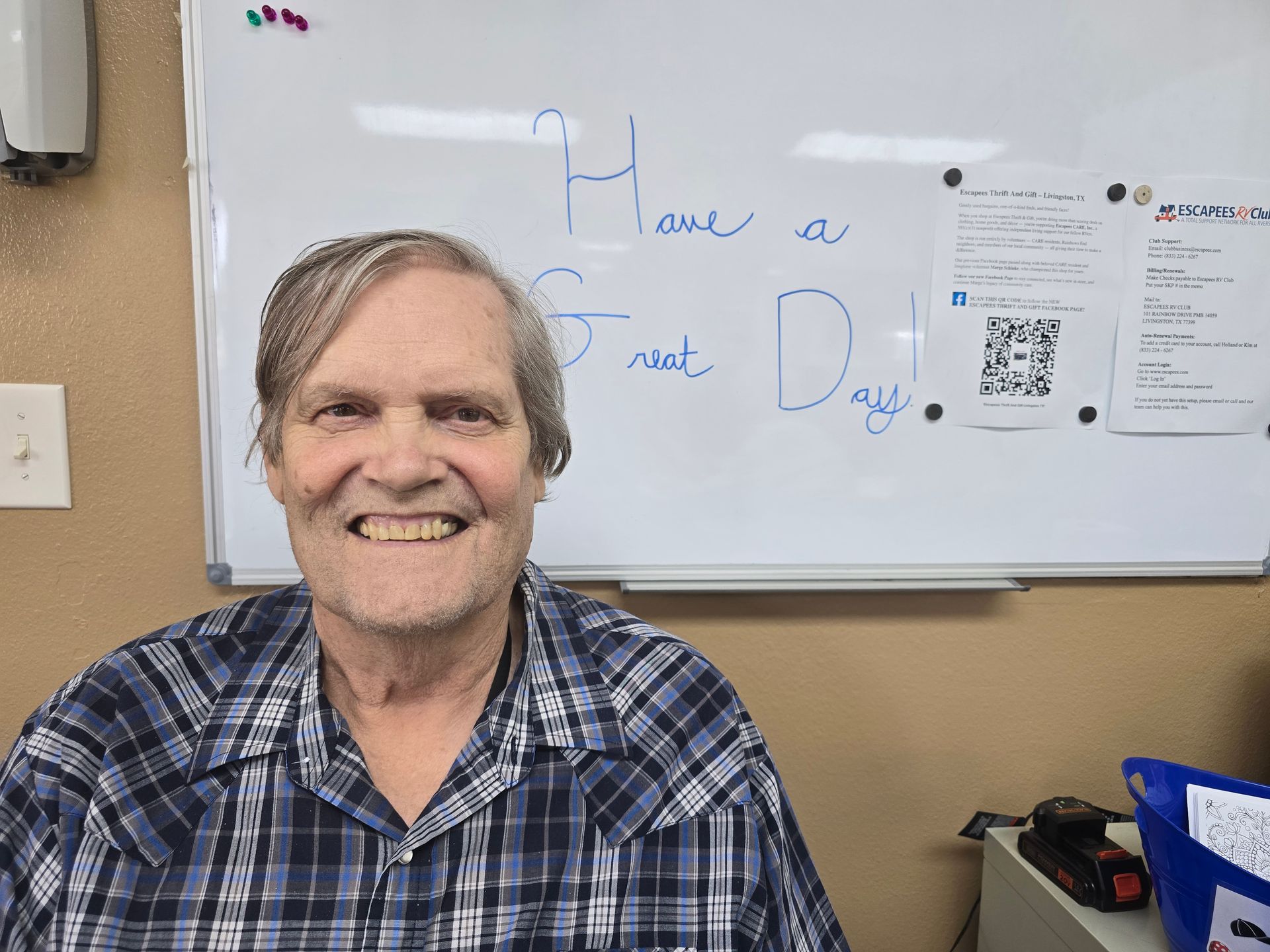 Smiling person in a plaid shirt seated in an office, with a whiteboard reading “Have a great day!”