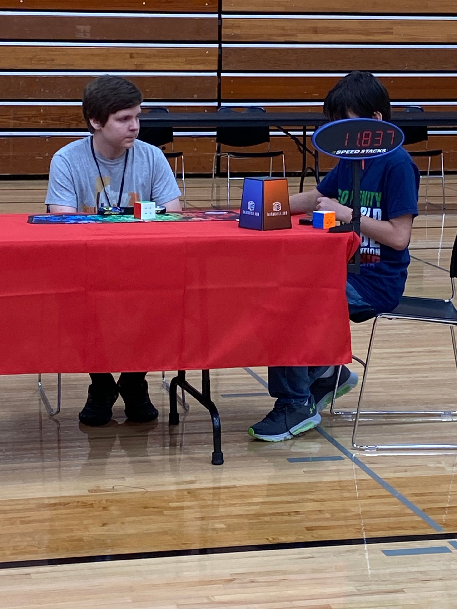 Two people sit at a red table in a gym, working with a model robot and parts during a competition.