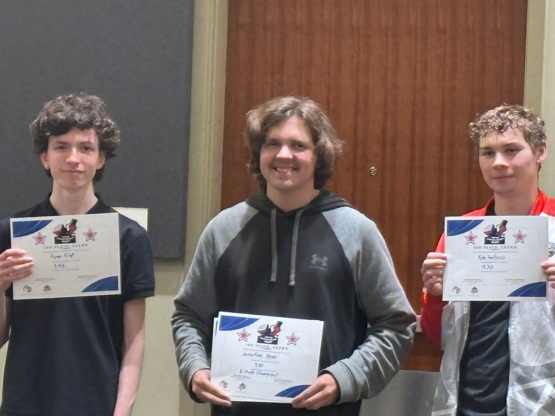 Three people holding certificates and smiling indoors in front of a wooden wall.