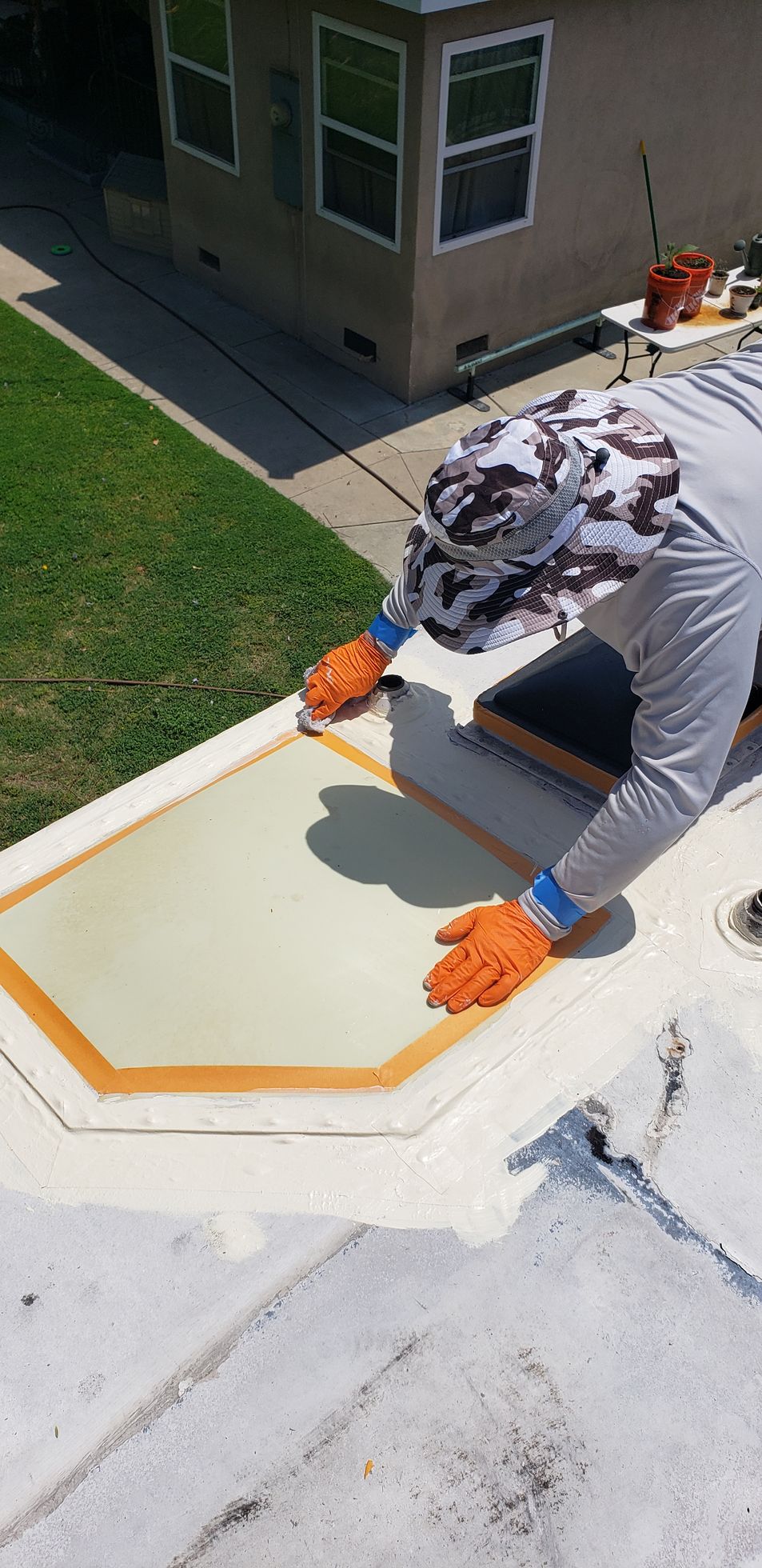 A person applying sealant to a skylight on a white roof, wearing gloves and a camouflage hat.