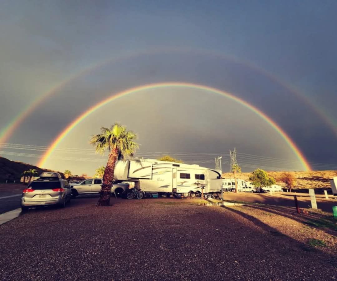Double rainbow arcs over a campsite with an RV, cars, and a palm tree.