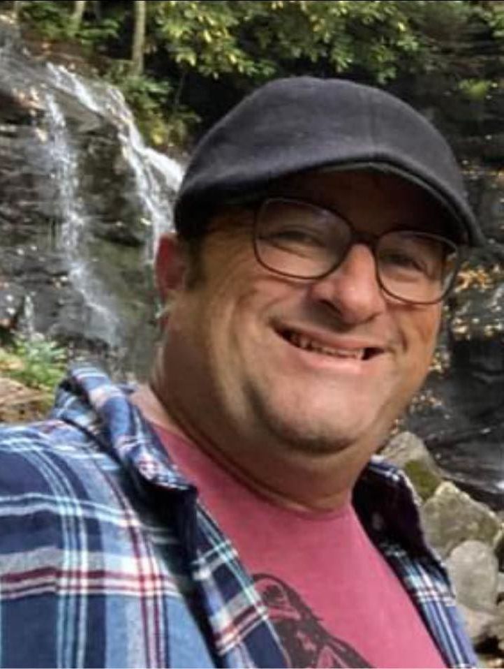 Man with glasses and hat smiles, wearing plaid shirt and red shirt, in front of a waterfall.