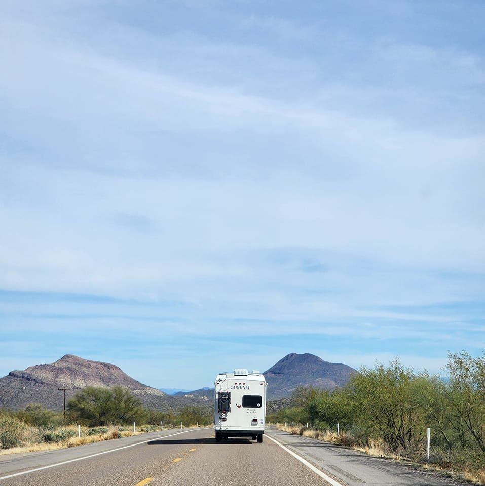 White RV driving on a desert highway with mountains in the background under a blue sky.