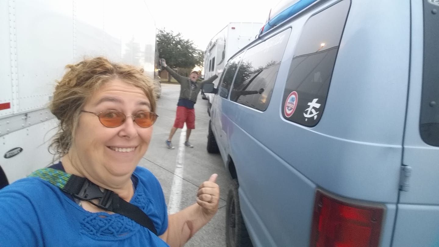 Woman gives thumbs up in front of a blue van, person in background with arms raised.