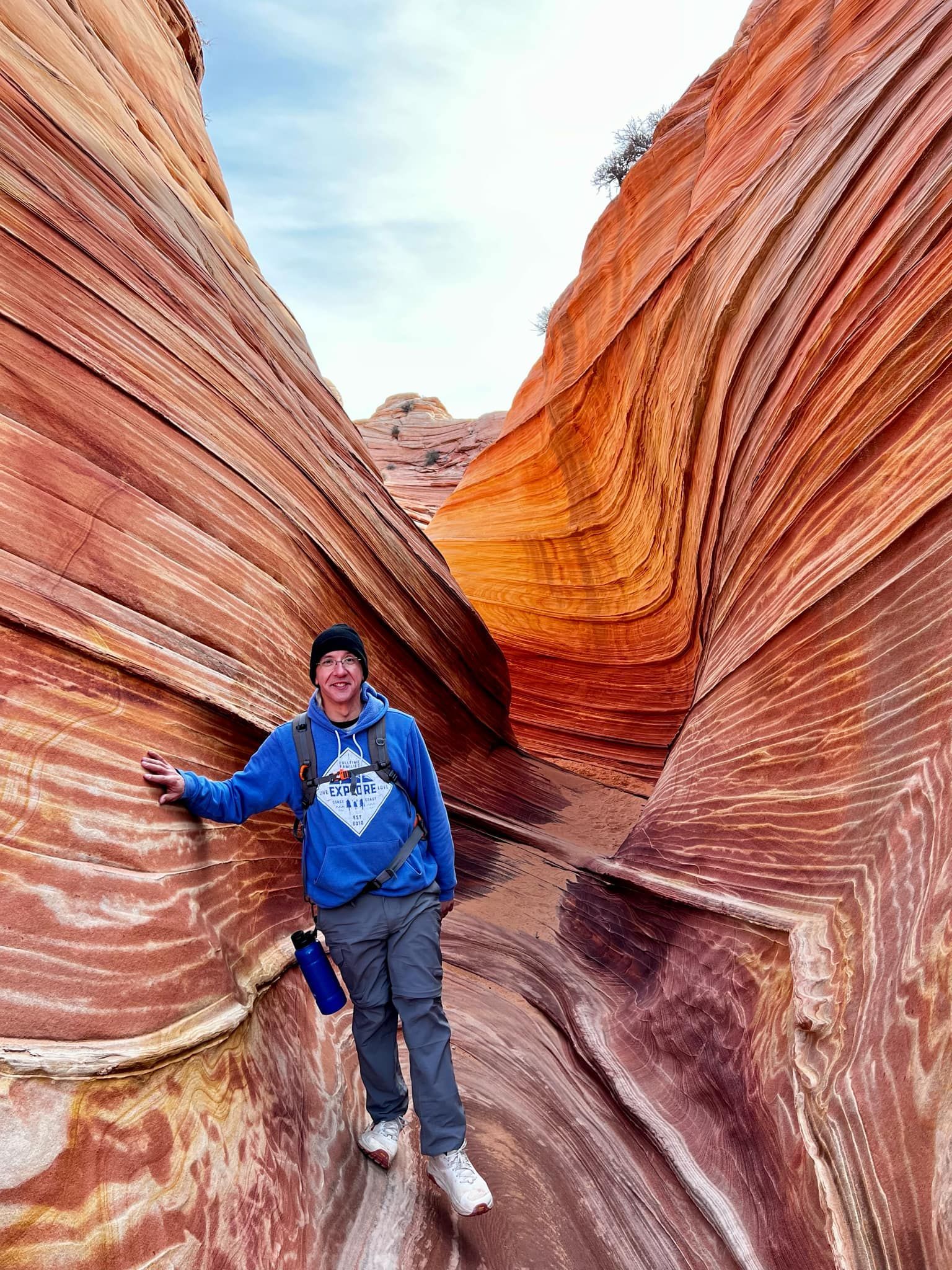 Man in blue sweatshirt and gray pants hiking in a narrow, wave-like sandstone canyon.