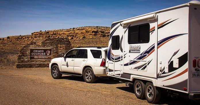 White SUV towing a camper trailer in front of a stone structure, brown landscape, sunny day.