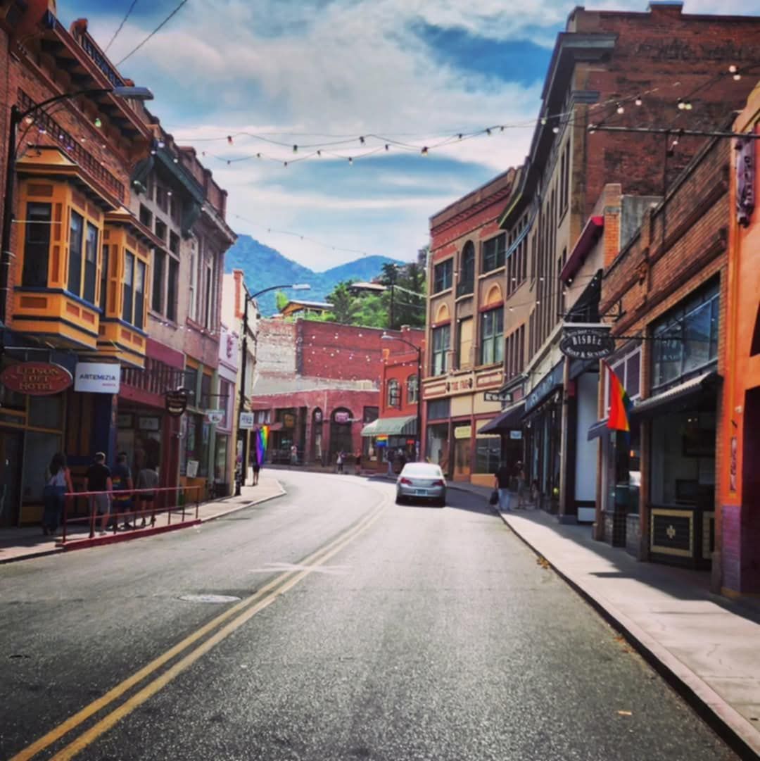 A street lined with brick buildings, a car, and string lights, with mountains in the background under a blue sky.