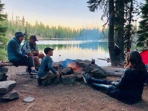 Family camping by a lake, roasting marshmallows over a fire at sunset.