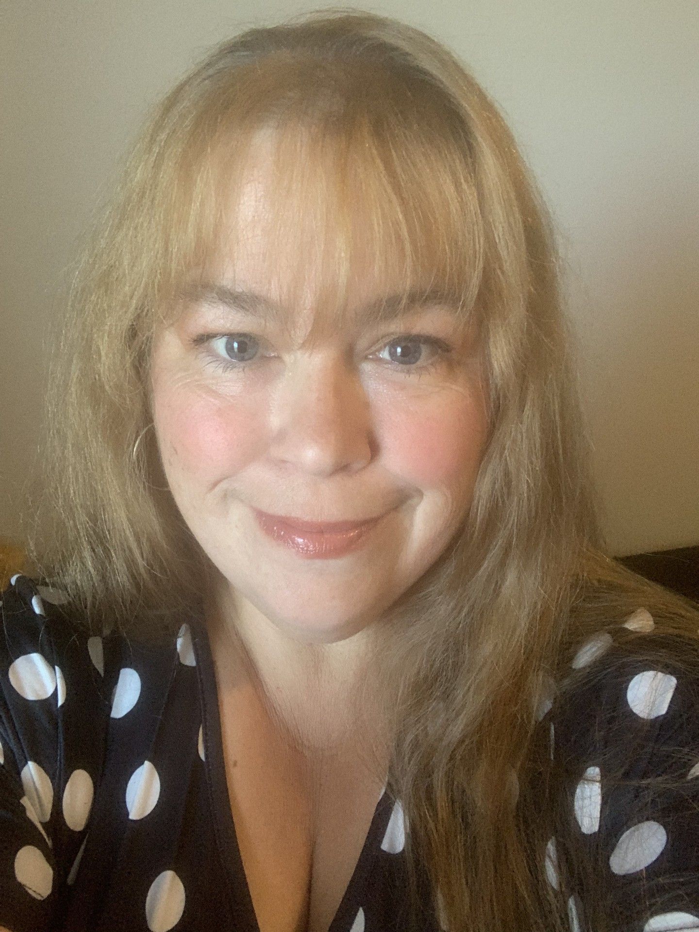 Woman with blonde hair and bangs, smiling, wearing a black and white polka-dot top.