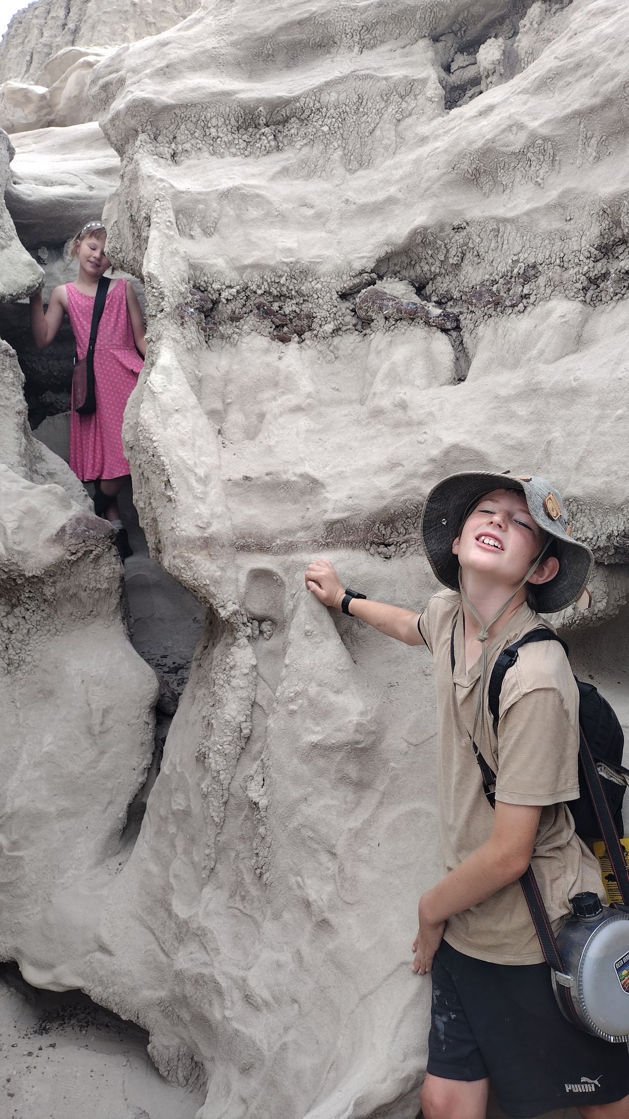 Two kids exploring a narrow, light-colored rock canyon. One is wearing a hat and backpack. The other is in a pink dress.