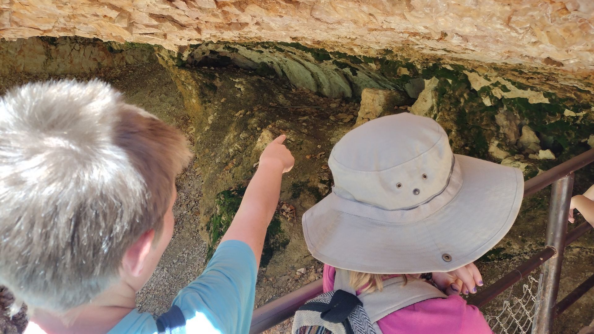 Two children, one pointing up, look at a green-hued cave wall. The other wears a wide-brimmed hat.