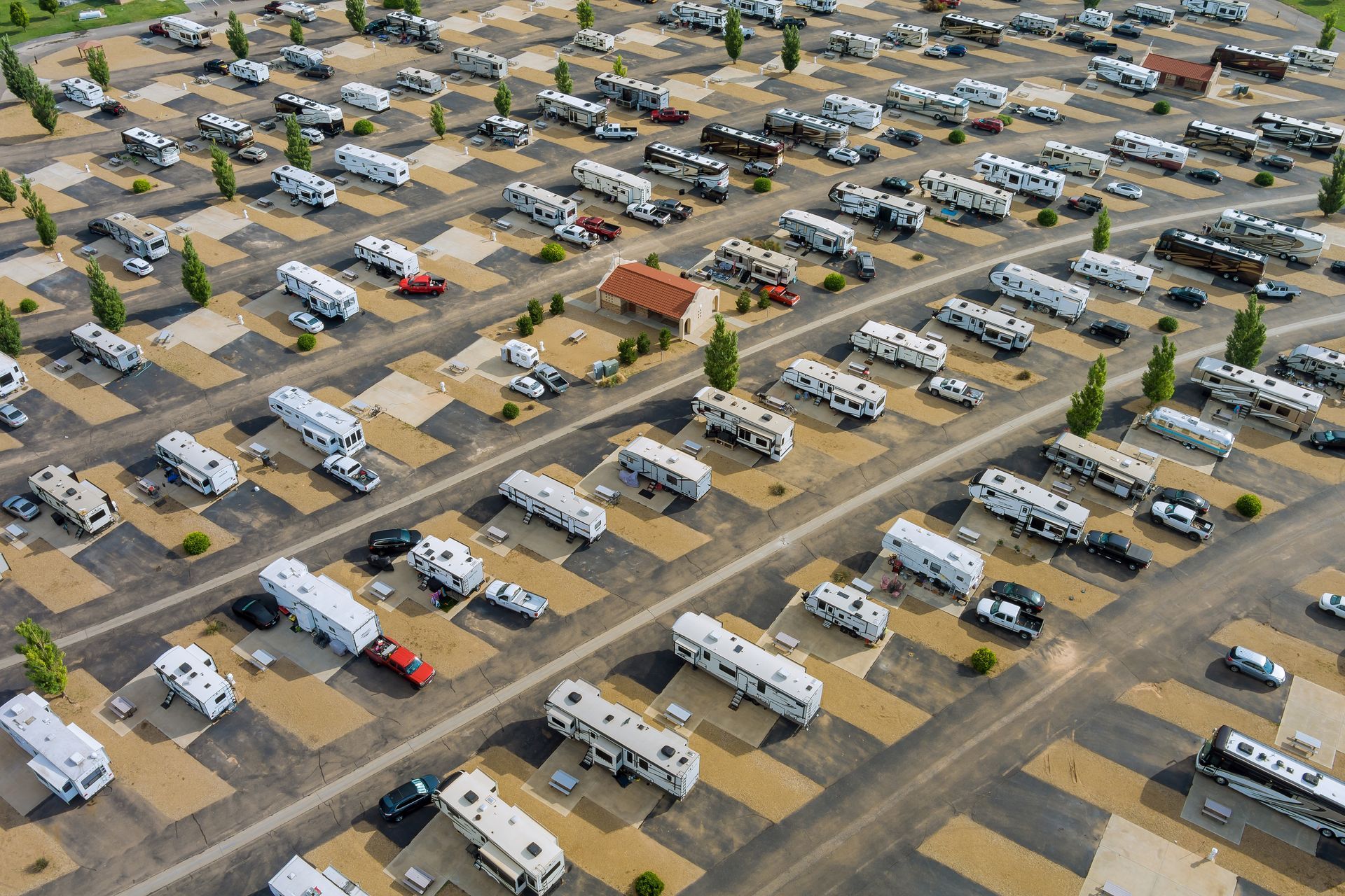 Aerial view of a packed RV park. Rows of RVs parked on gravel sites. Trees and a small building are interspersed.