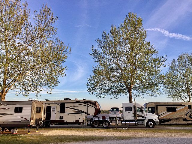 RVs parked in a row, with a semi-truck in between, trees, and a partly cloudy sky.