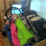 A cluttered RV interior with a stroller, bags, and other items piled up near a window.