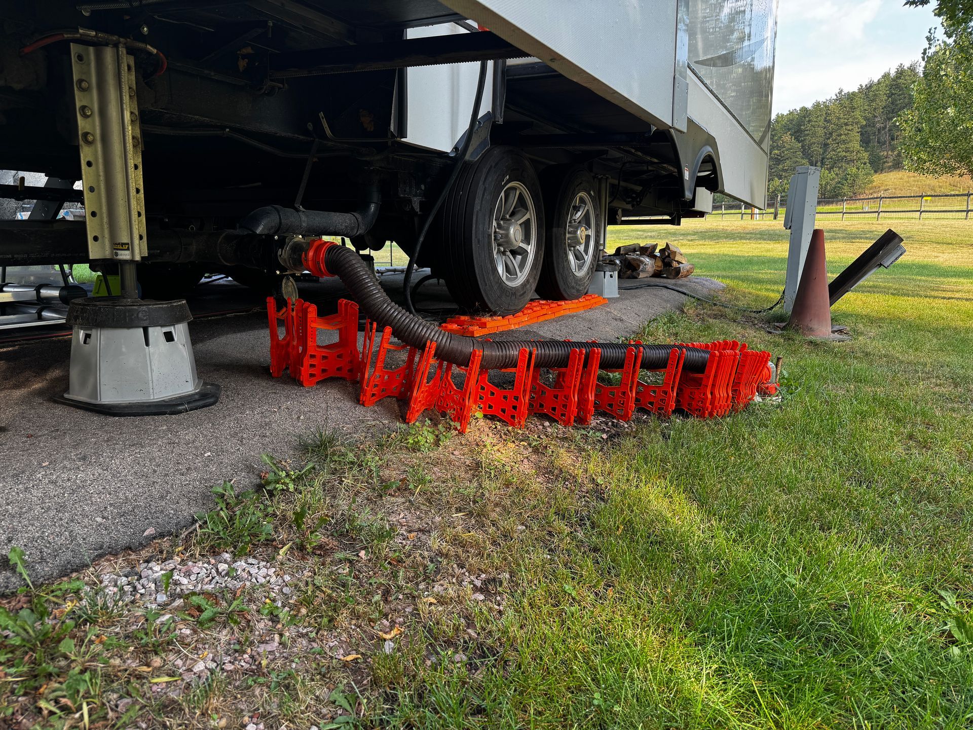 RV with orange hose supports connected to a drain. Black tires on gravel, green grass and a wooded area.