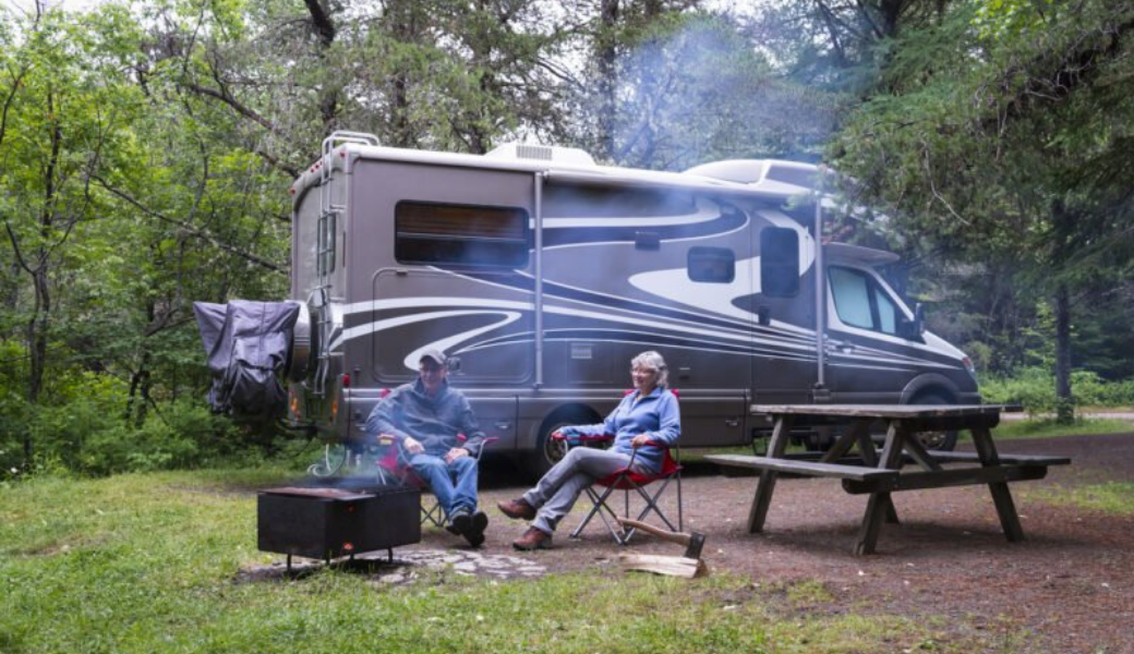 Couple camping by an RV, sitting near a fire pit and picnic table. Smoke rises.