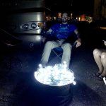 Man seated with a tray of lit lights. Night scene, street.