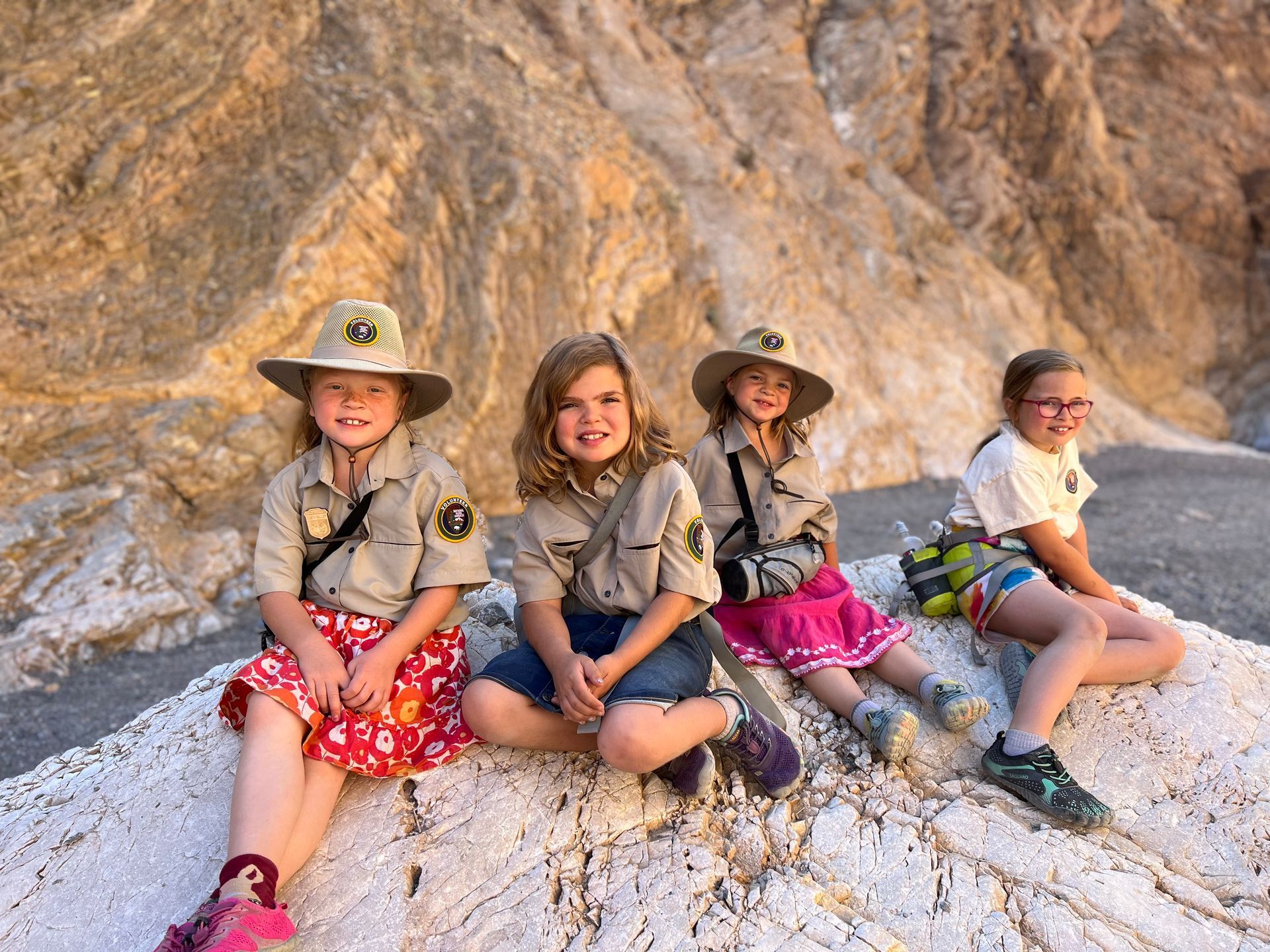 Four young girls wearing scout uniforms and hats sit on a light-colored rock in a canyon.