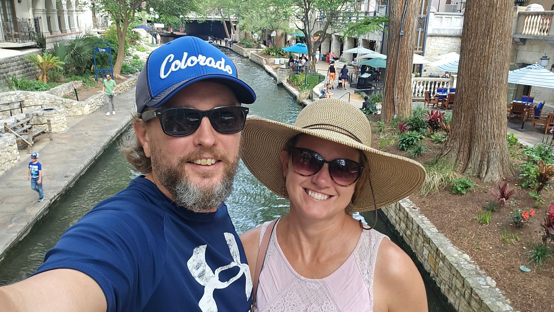 Woman with glasses smiles with hat in front of a canal next to man in Blue colorado hat