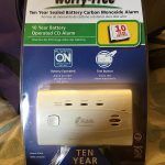 A package of a carbon monoxide alarm is sitting on a bed.