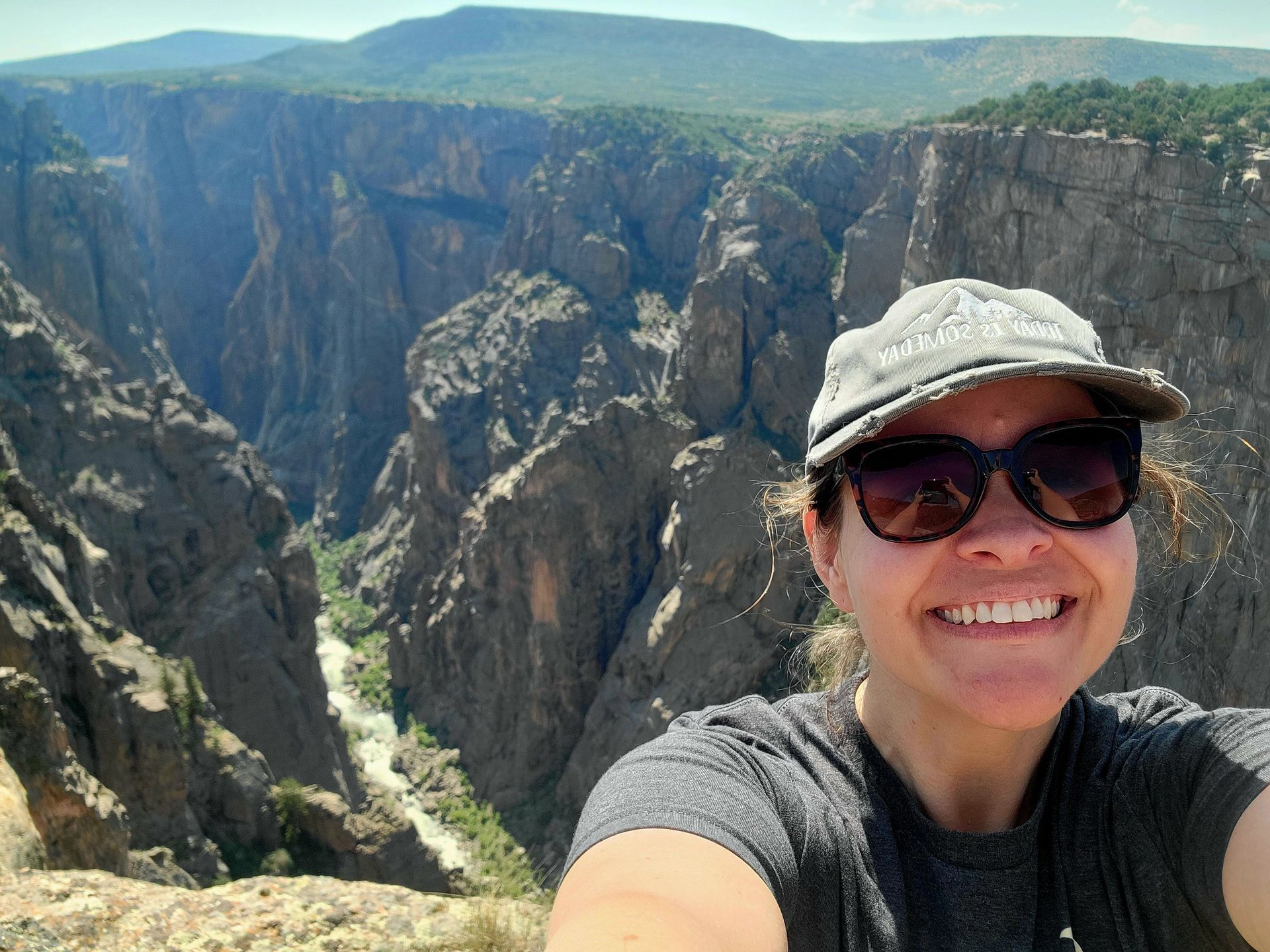 A woman wearing sunglasses and a hat is taking a selfie in front of a canyon.