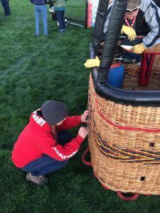 Person in red hoodie working on the wicker basket of a hot air balloon on a grassy field.