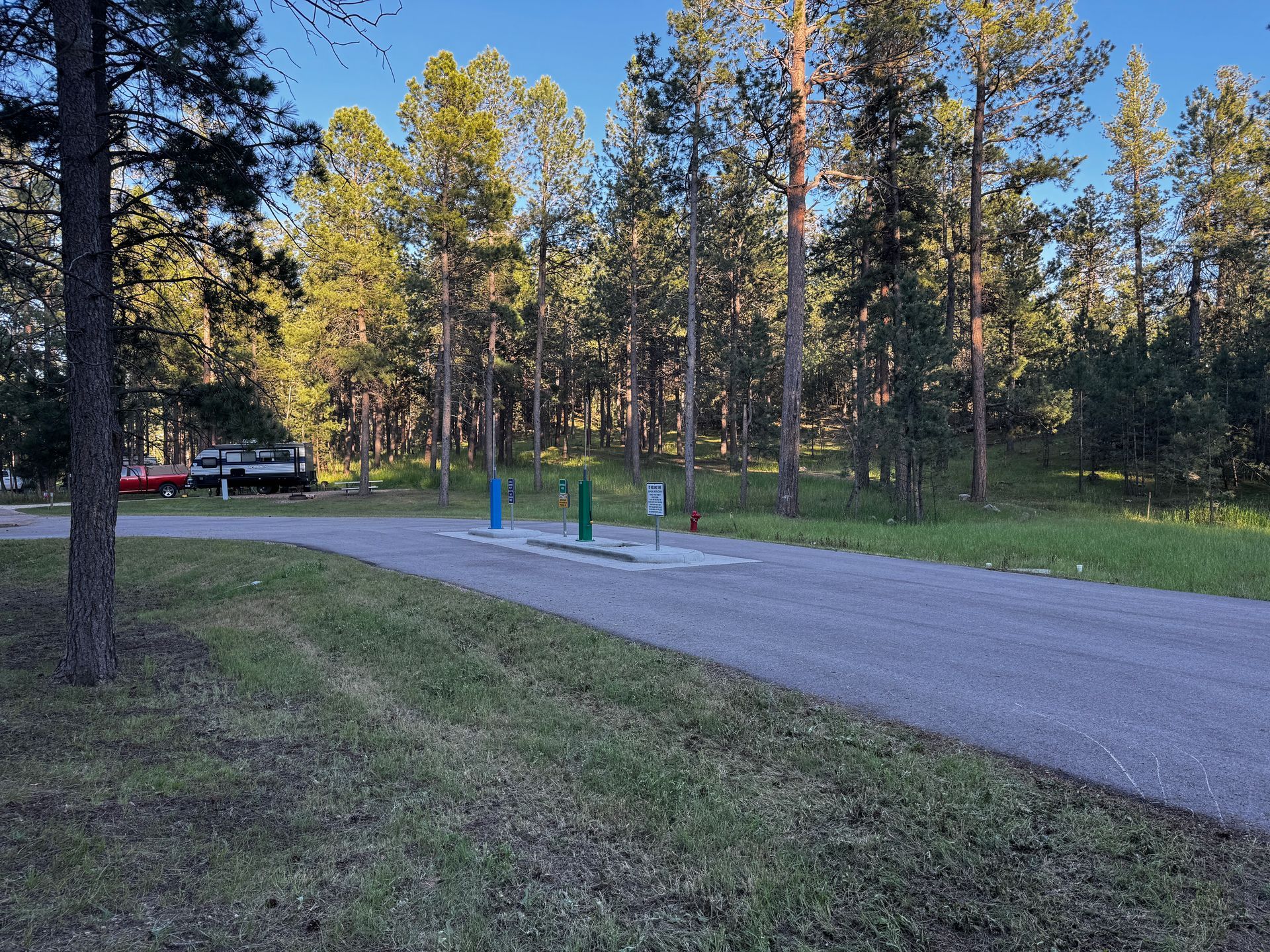 Campsite with asphalt road, water hookups, and trees. Vehicles parked in the distance.