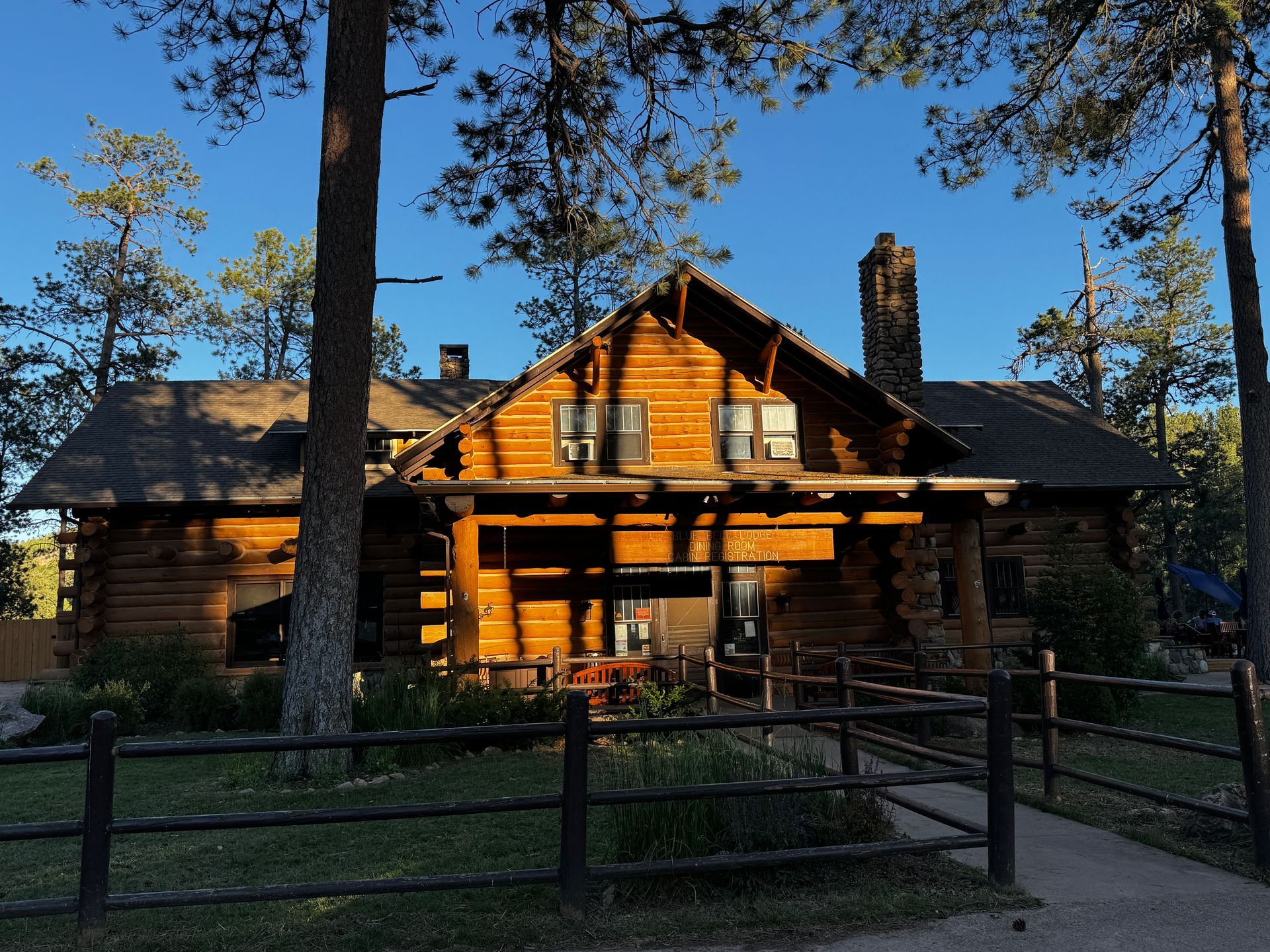 Log cabin, wooden, with a chimney, surrounded by trees and a fence.