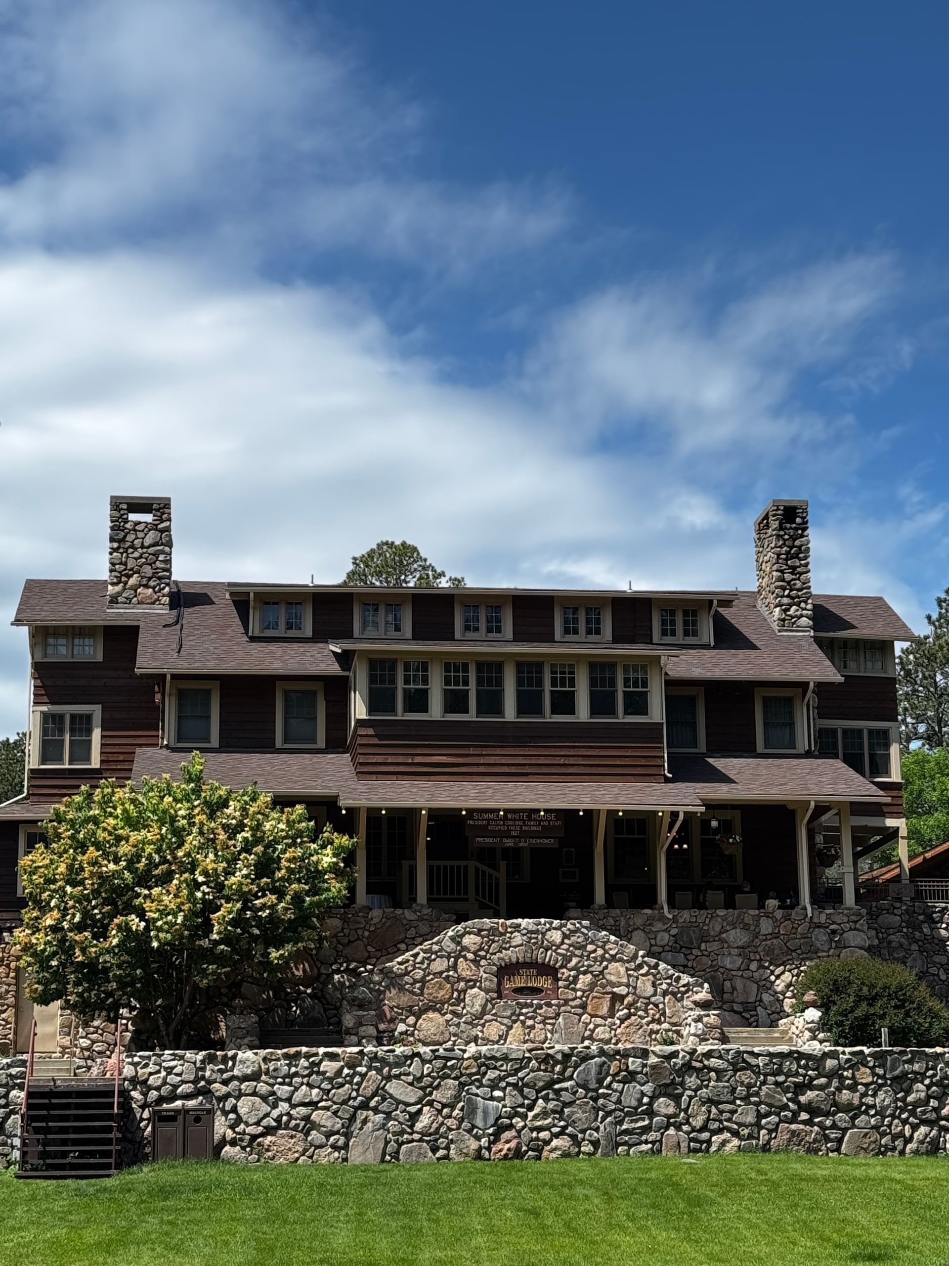 Large brown building with stone chimneys, built into a hill, with stone wall and green lawn below, blue sky.