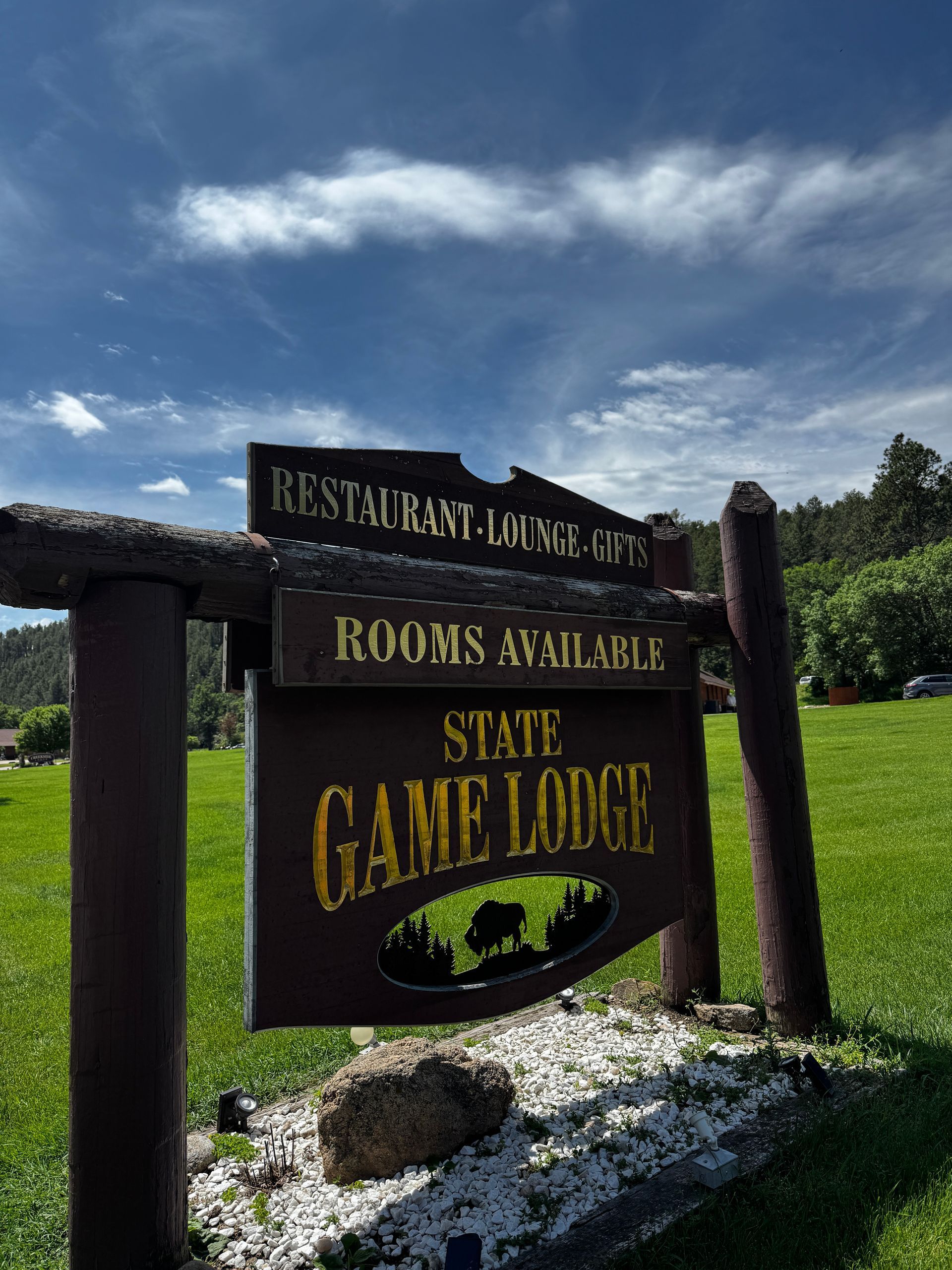 Sign for State Game Lodge, brown with gold lettering, indicating rooms, restaurant, and gift shop, set on green lawn.