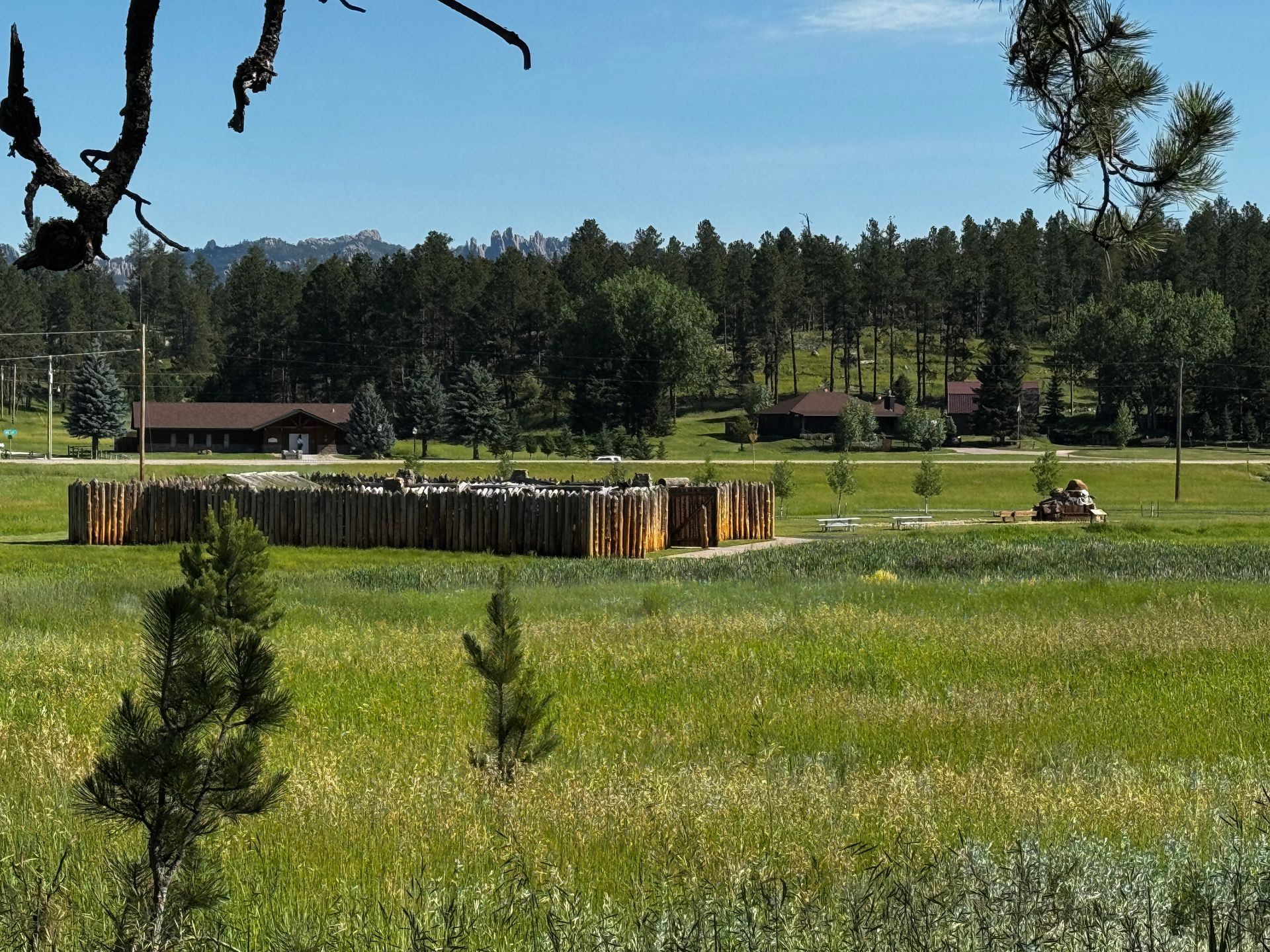 A wooden stockade in a grassy field, surrounded by trees, under a blue sky.