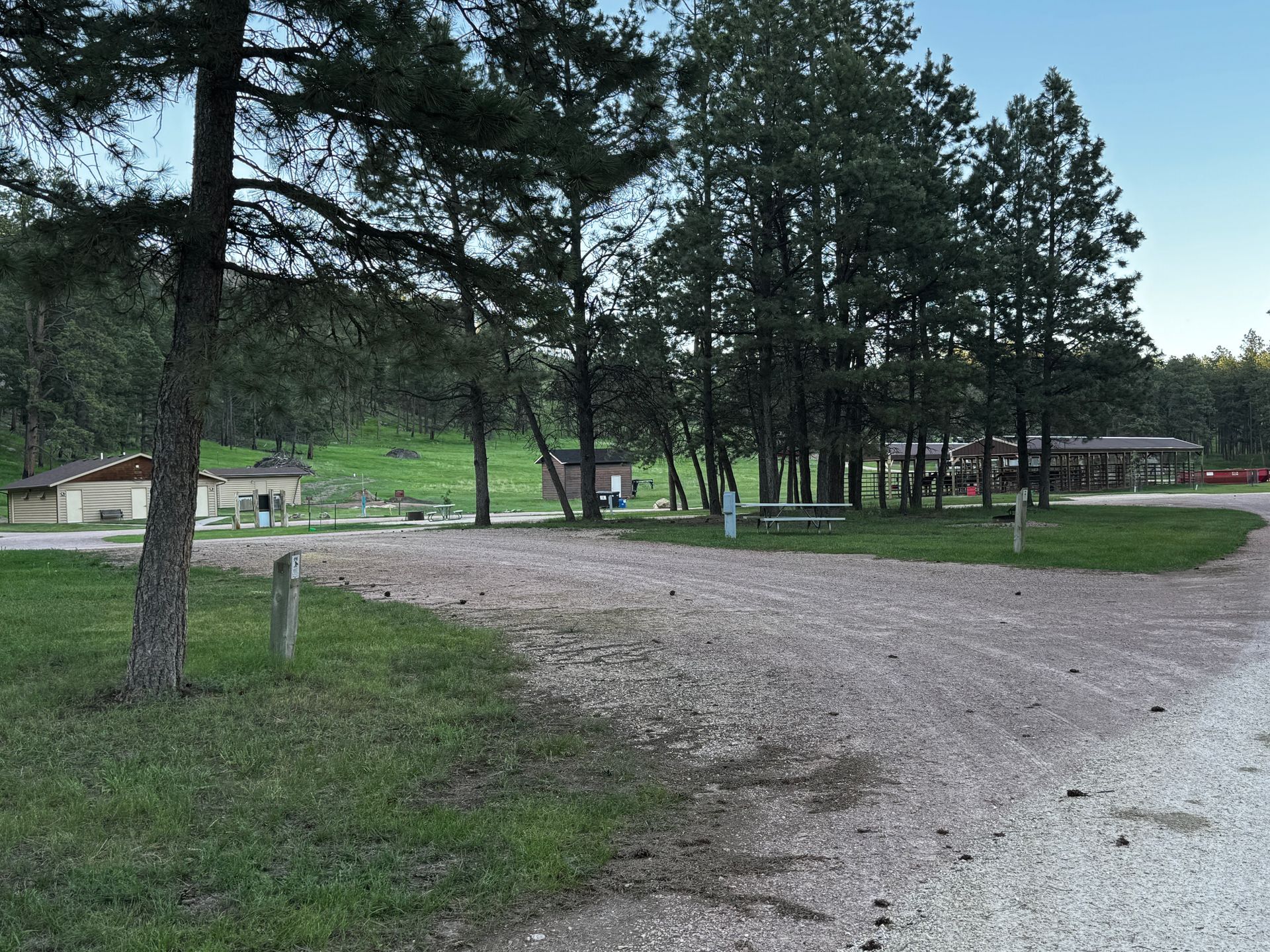 Gravel parking area in a grassy field with trees and restrooms in a park setting.
