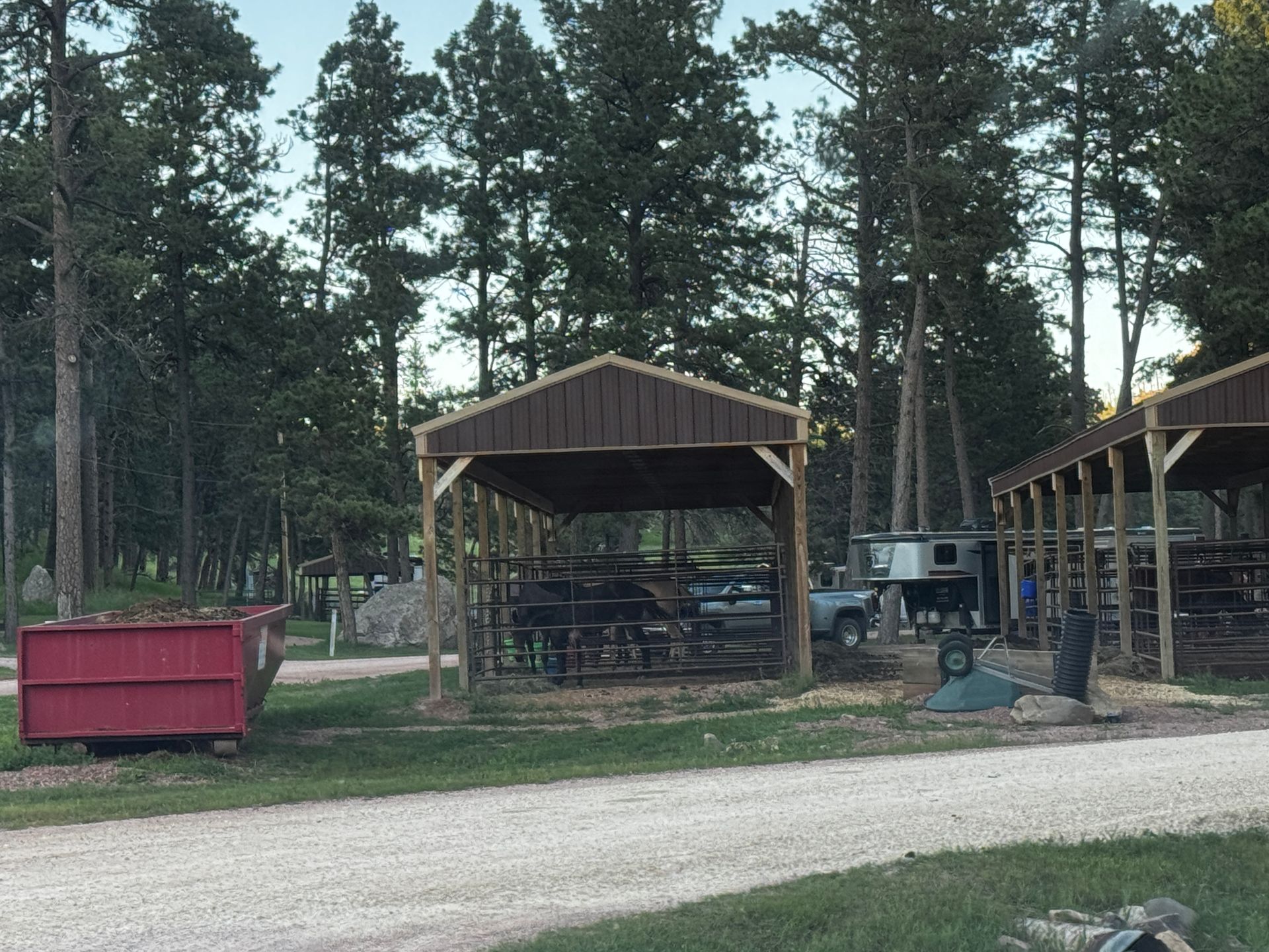 Camping area with covered shelters, trees, and a red dumpster.