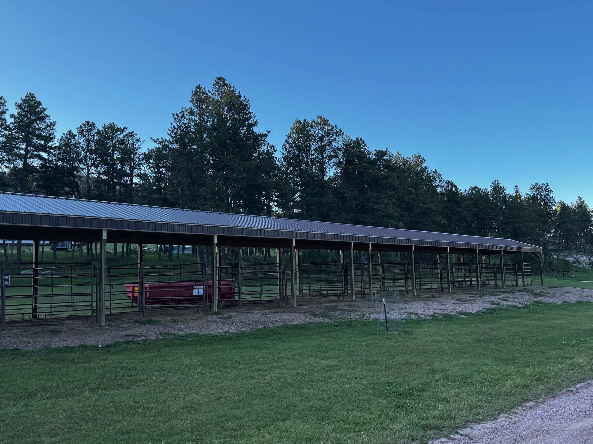 A long, open-sided shed with a metal roof in front of a line of trees.