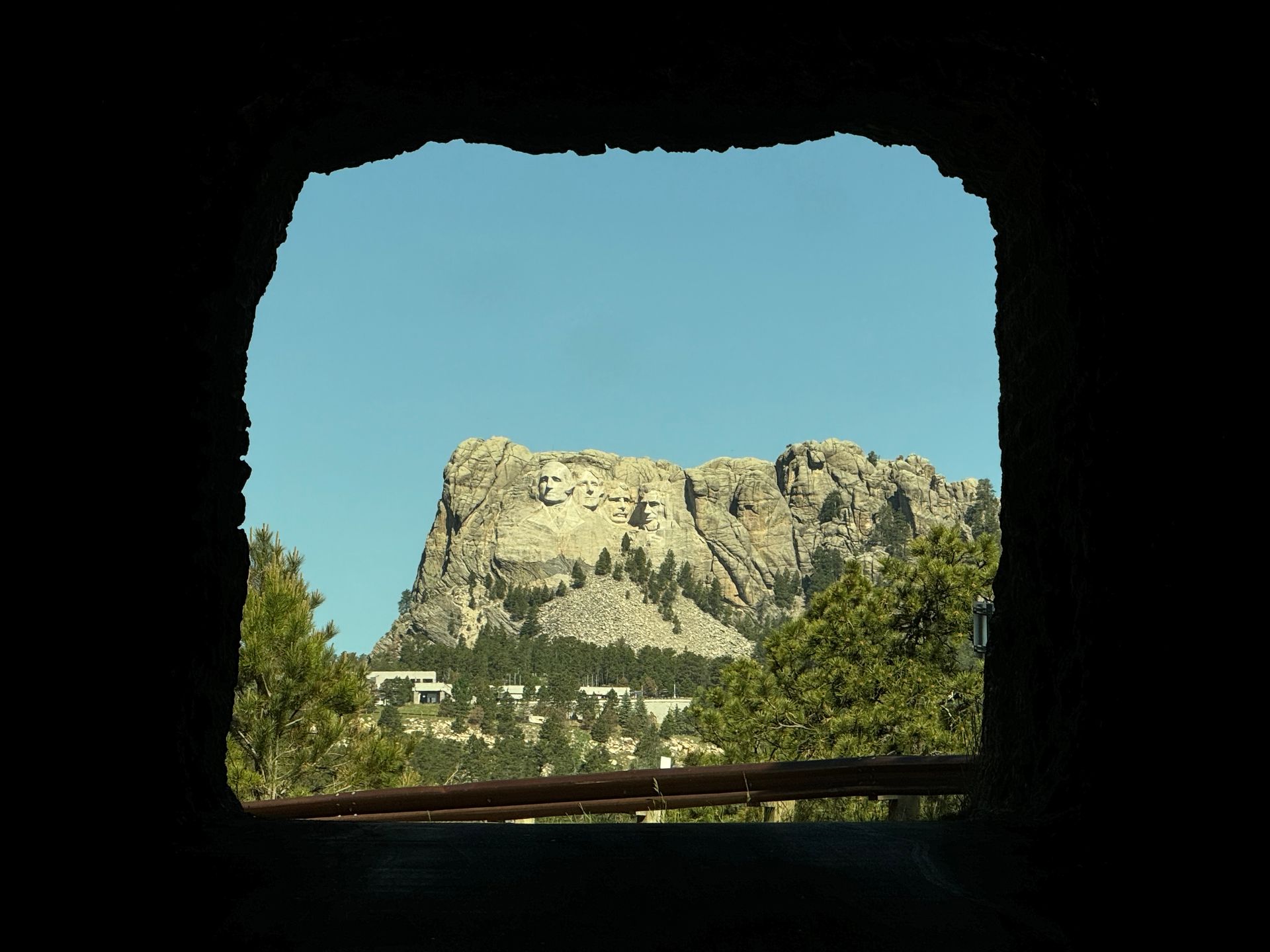Mount Rushmore framed by a dark tunnel opening; blue sky, trees, and a small building are visible.