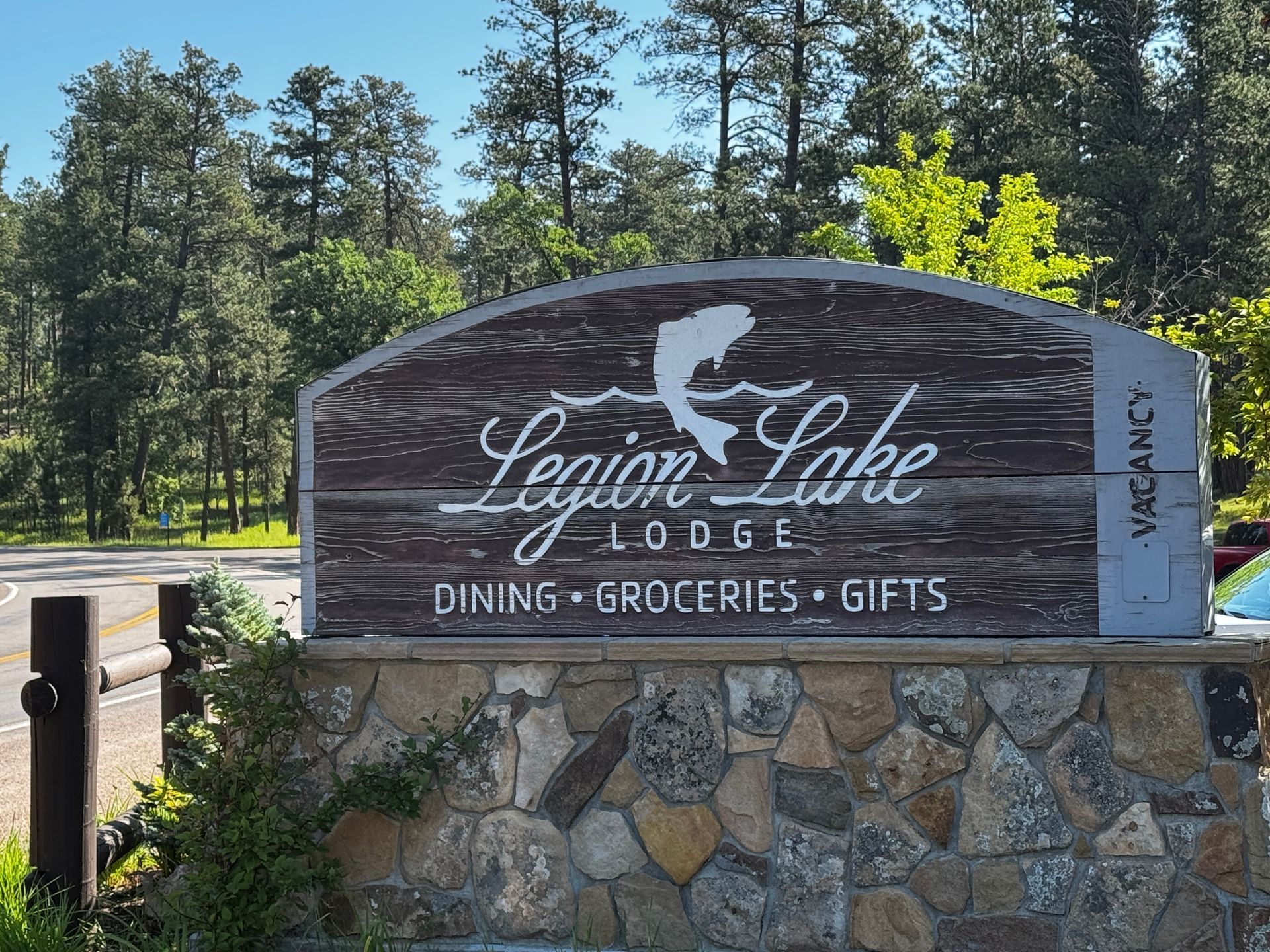 Sign for Legion Lake Lodge: Dining, Groceries, Gifts. Stone base, brown wooden sign with a fish silhouette.
