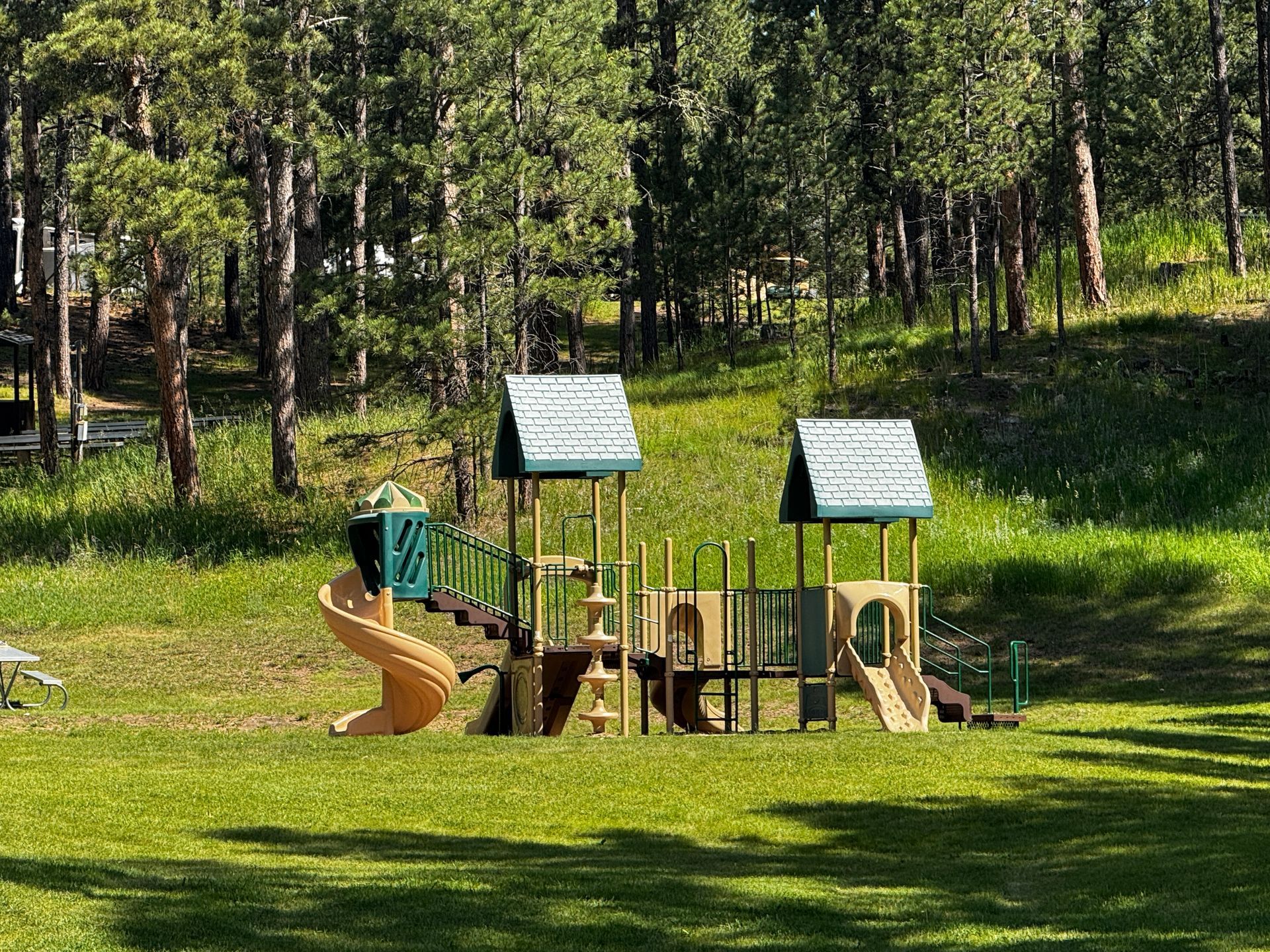 Playground in grassy area, with a tan slide, brown structure, and green roofs. Surrounded by trees.